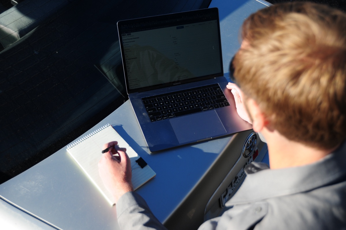 Officer writing notes in a spiral notepad with a laptop on a vehicle hood